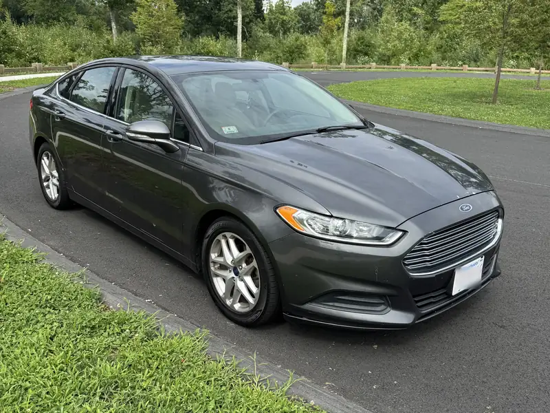 Grey Ford Fusion sedan with tan interior, front three-quarter view, parked on a tree-lined road